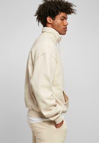 Beige genser med høy krage, oversized passform og kengurulomme. Myk tekstur med ribbestrikkede mansjetter og søm. Sammen med matchende bukser.