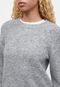 Person with curly blonde hair wearing a soft, light grey knit sweater over a white crew neck shirt.