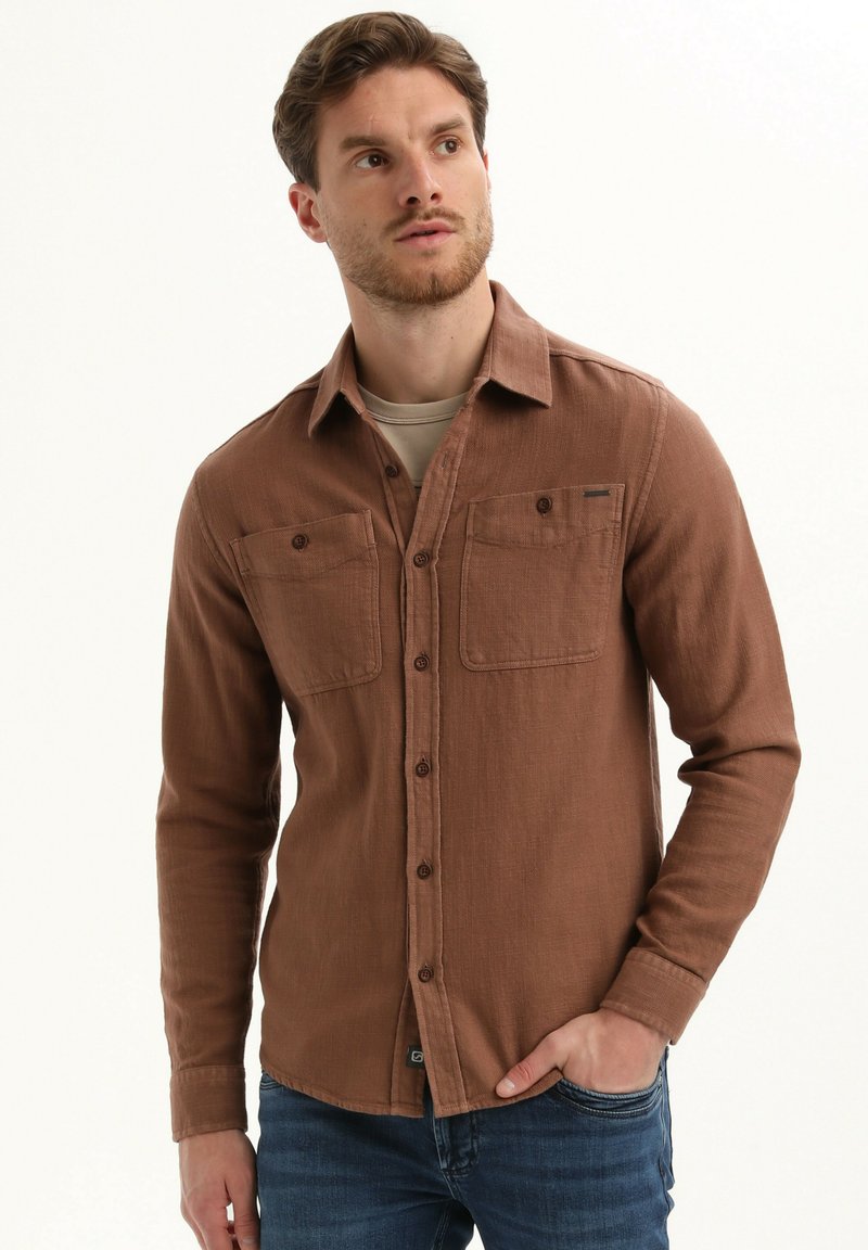 Rjava srajca s gumbi iz teksturirane tkanine, z dvema prsnama žepoma, dolgimi rokavi in klasičnim ovratnikom. Uporabljena z modrimi kavbojkami.
