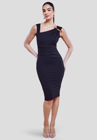 Donna che indossa un vestito nero aderente a spalla unica con una fibbia decorativa, in piedi con le mani sui fianchi contro uno sfondo chiaro e uniforme.