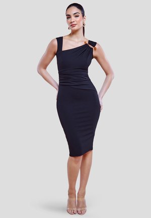 Donna che indossa un vestito nero aderente a spalla unica con una fibbia decorativa, in piedi con le mani sui fianchi contro uno sfondo chiaro e uniforme.