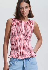 Red and white checkered sleeveless top with smocked detailing, round neckline, and a ruffled hem, paired with light blue denim jeans.