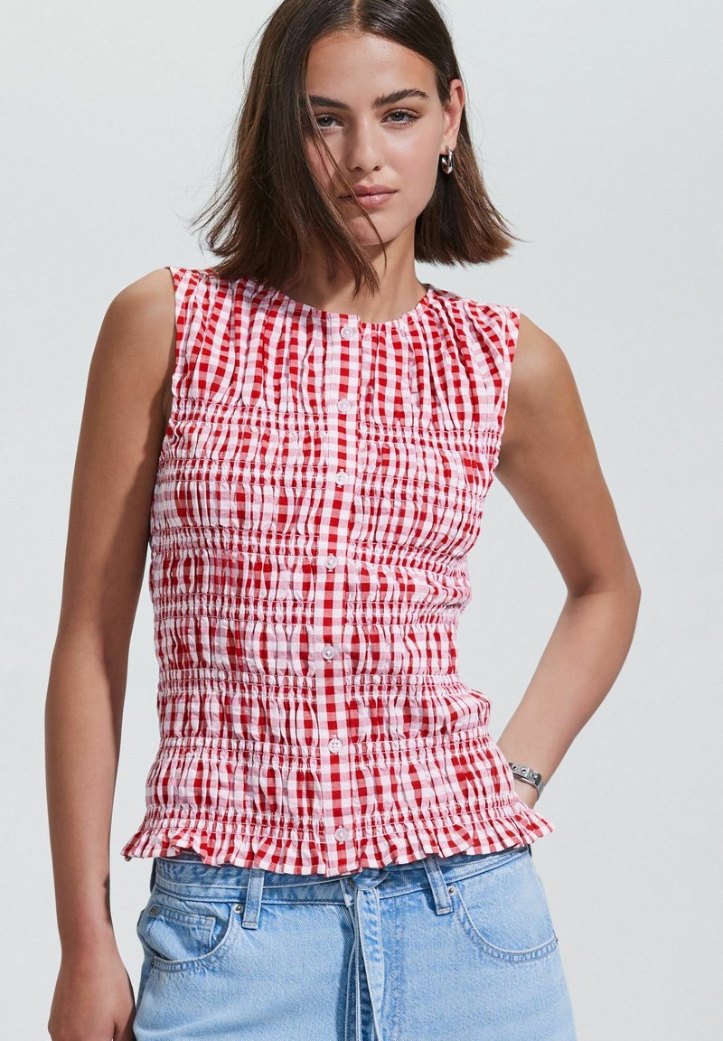 Red and white checkered sleeveless top with smocked detailing, round neckline, and a ruffled hem, paired with light blue denim jeans.
