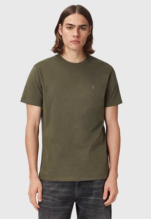 Ung mand med skulderlangt brunt hår iført en olivengrøn t-shirt med korte ærmer og falmede sorte jeans, stående foran en ensfarvet baggrund.