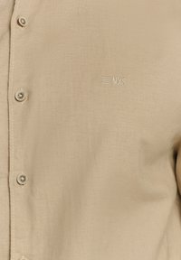 Beige knoopshirt gemaakt van textiel met een gestructureerde stof, met een klein geborduurd logo op de borst en contrasterende knopen.