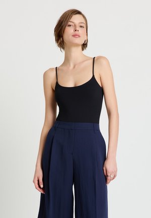 Donna con capelli corti che indossa un top nero a spalline sottili e pantaloni blu navy a gamba larga e vita alta, in piedi davanti a uno sfondo chiaro.