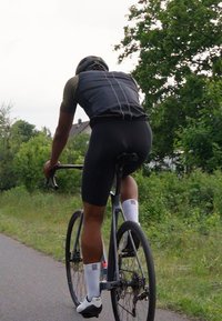 Un cycliste vêtu d'une tenue aérodynamique noire avec des accents réfléchissants roule sur un vélo argenté élégant sur un chemin, entouré de feuillage vert.