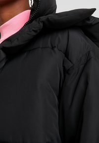 Svart vadderad jacka med hög krage, med en slät yta och synliga sömmar. En rosa tröja med rund hals är delvis synlig under.