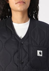 Schwarze wattierte Weste mit V-Ausschnitt, Knopfverschluss und aufgesetzter Tasche mit Logo. Glatte Textur und dezentes Wellenmuster sichtbar.