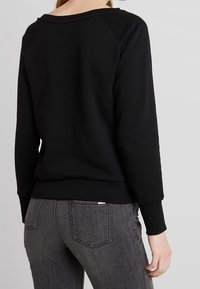 Felpa nera con scollatura ampia e maniche raglan, caratterizzata da polsini e orlo a coste. Indossata con jeans grigio scuro. Tessuto liscio.