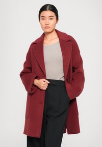 Manteau en laine bourgogne avec un col cranté, coupe oversized et fermeture à bouton unique. Associé à un haut gris à côtes et un pantalon à rayures.