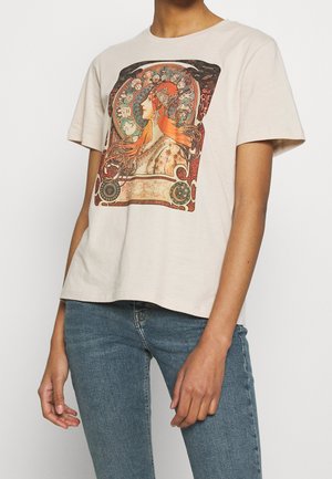 Person iført beige kortærmet skjorte med farverigt vintage kunstprint af kvinde med flagrende hår, kombineret med blå jeans.
