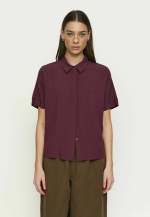 Donna dai capelli castani lisci che indossa una camicia bordeaux a maniche corte e pantaloni verde oliva, in piedi contro uno sfondo neutro.