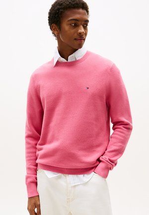 Rosa Strickpullover mit einem strukturierten Design, Rundhalsausschnitt, gerippten Bündchen und Saum. Logo-Patch auf der Brust. Kombiniert mit einem weißen Hemd mit Kragen.