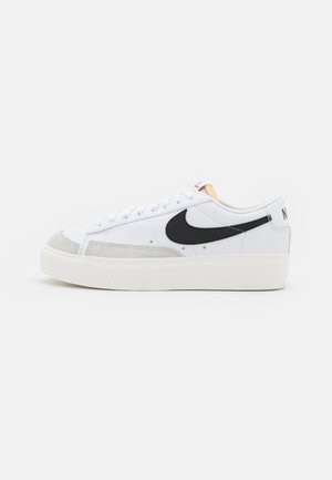 Witte lage sneaker met zwarte Nike-swoosh, lichtgrijze suède neuskap, witte veters en dikke off-white zool op een effen achtergrond.
