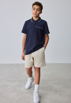 Adolescent marchant, portant un polo bleu marine à manches courtes, un short beige, des chaussettes blanches et des baskets blanches, sur un fond gris clair uni.