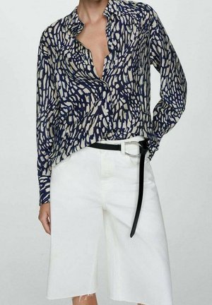 Persoon draagt een marineblauw-wit gemerkt overhemd, ingestopt in hooggetailleerde witte denimshorts met een loshangende zwarte riem.