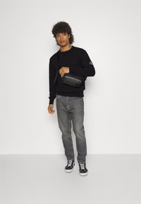 Felpa nera, jeans grigi e sneakers neri con suole bianche. Il modello tiene una piccola borsa a tracolla nera. Design semplice e colori solidi.