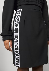 Svart kjol med vit vertikal rand på sidan som har texten "VERSACE JEANS COUTURE", bärs med en svart långärmad topp, hand synlig.