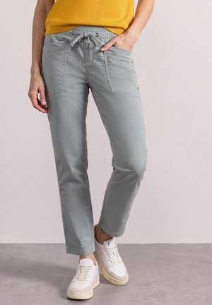 Personne portant un pantalon court gris clair avec des poches à l'avant, un haut jaune et des baskets blanches, debout sur une surface grise.