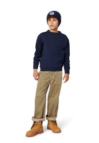 Pull en maille bleu marine, pantalon en velours côtelé beige et bottes à lacets tan avec des lacets jaunes. Le pull présente une manche texturée et un écusson logo.