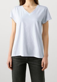 Donna che indossa una maglietta a maniche corte blu chiaro con scollatura a V e jeans neri, in piedi contro uno sfondo semplice chiaro.