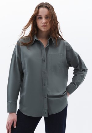 Grå skjorte med knapper lavet af glat stof, med en brystlomme, lange ærmer og en buet kant. Bæres med mørkeblå jeans.