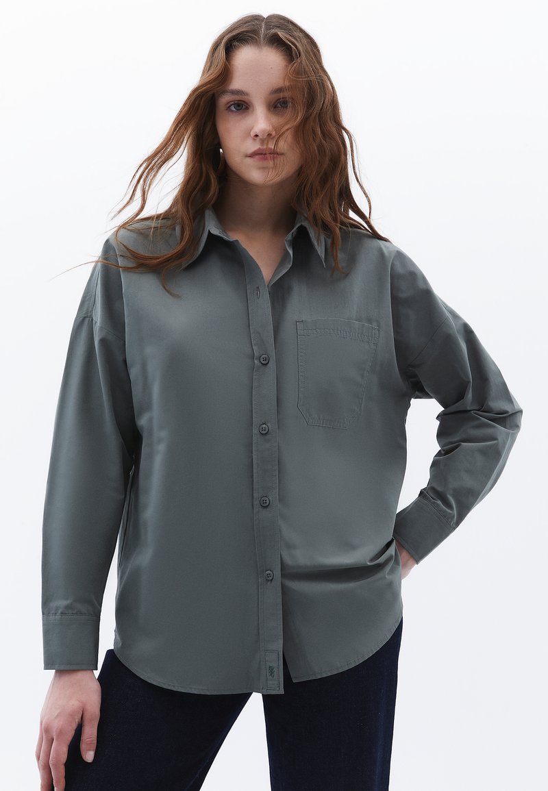 Camicia grigia con bottoni realizzata in tessuto morbido, con taschino frontale, maniche lunghe e orlo curvo. Indossata con jeans blu scuro.
