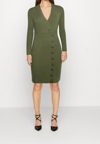 Femme portant une robe portefeuille côtelée olive verte jusqu'aux genoux, à manches longues et boutons noirs sur le côté gauche, associée à des talons noirs à brides.