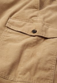 Tissu en velours côtelé marron clair avec une texture douce, présentant une poche poitrine avec un bouton foncé et des détails cousus.