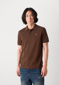 Braunes Poloshirt mit kurzen Ärmeln und Kragen, das mit einem kleinen grünen Logo versehen ist. Der Stoff wirkt glatt; getragen mit blauen, ausgewaschenen Jeans.