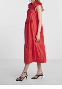 Robe midi en coton rouge avec des manches volantées, un devant boutonné et des détails brodés à l'ourlet. Portée avec des talons strappy noirs.