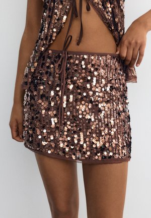 Personne portant une mini-jupe marron à sequins avec un haut assorti, tous deux ornés de sequins ronds brillants et de détails à cordon, se tenant devant un fond uni.