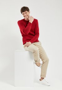 Der rote Strickpullover hat einen Rundhalsausschnitt und Schulterknopf-Akzente, kombiniert mit beigen Chinos und weißen Sneakers, die auf einem weißen Podest sitzen.