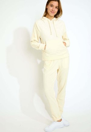 Cremefarbener Hoodie und Jogginghosen-Set, ausgestattet mit einer Kängurutasche, verstellbarem Kapuzenzug und gerippten Bündchen, kombiniert mit weißen Sneakers.