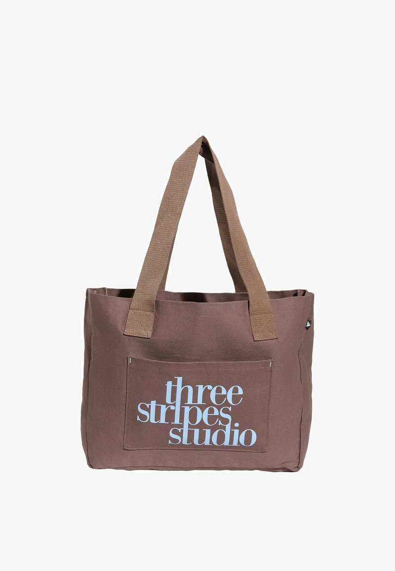 Brown fabric tote bag with wide handles and front pocket displaying "three stripes studio" in light blue text.