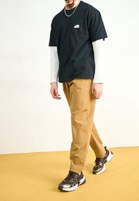 Černé tričko s krátkým rukávem a kapsou; béžové cargo kalhoty; tmavé tenisky s bílými akcenty; stojící na žlutém koberci.