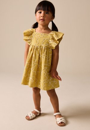 Ung pige iført gul kjole med hvide blomsterprint og hvide sandaler stående mod en ensfarvet beige baggrund.