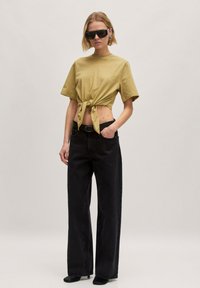 Khaki cropped t-shirt met een knoopdetail aan de voorkant, gecombineerd met hoge zwarte jeans en zwarte schoenen. Minimalistische zonnebril en accessoires.