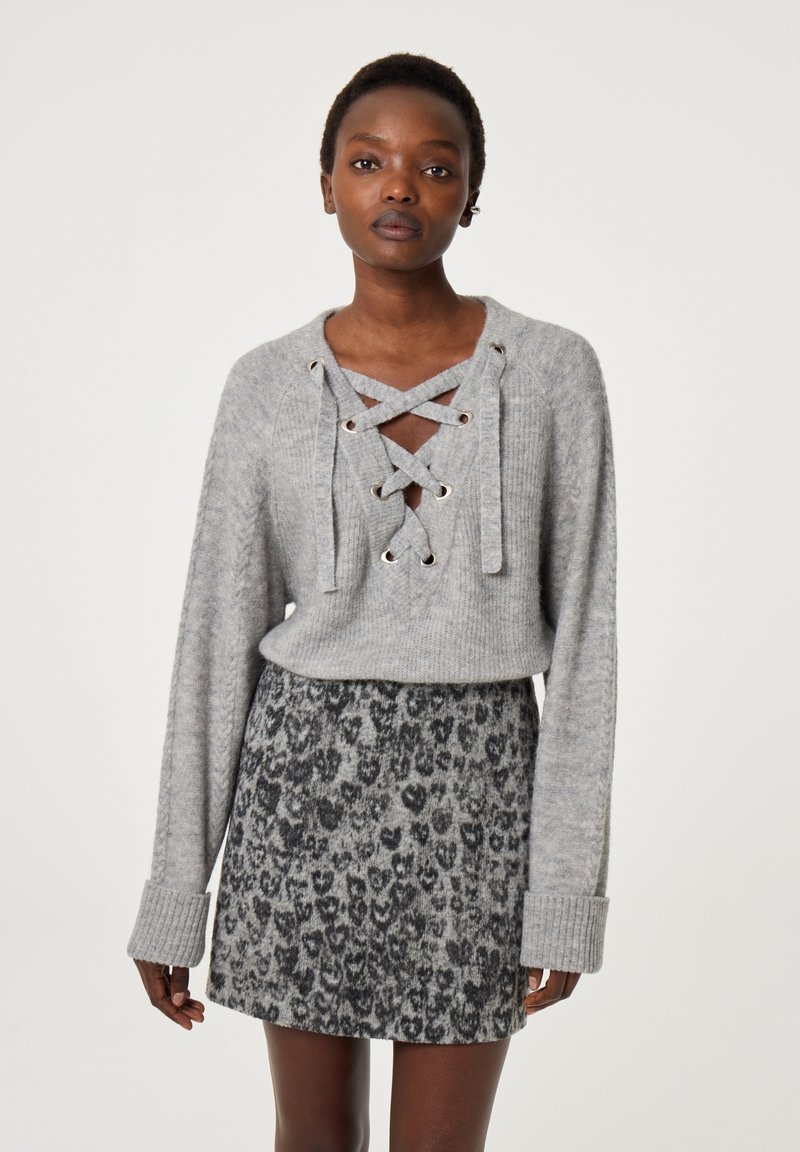 Grauer Strickpullover mit Schnürdesign und metallischen Ösen, kombiniert mit einem kurzen Rock im grau-schwarzen Leopardenmuster.