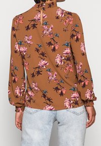 Braune Bluse mit langen Ärmeln und floralem Muster. Sie hat einen hohen Kragen mit rüschenförmigem Saum und rosa sowie blauen Blumenmustern. Knopfverschluss im Rücken.