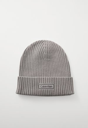 Bonnet tricot gris avec une texture côtelée, présentant un revers replié et une étiquette logo rectangulaire avec "Calvin Klein" en blanc.