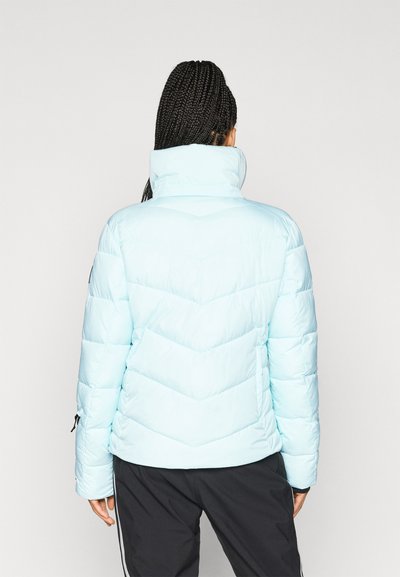 Ανοιχτού μπλε φουσκωτό μπουφάν με ψηλό γιακά, ραφές σε σχήμα V, ελαστικές μανσέτες και χαλαρή εφαρμογή, συνδυασμένο με μαύρο παντελόνι.