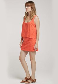 Débardeur corail avec broderie florale et ourlet festonné, associé à une jupe assortie. Porté avec des sandales brunes à lanières.