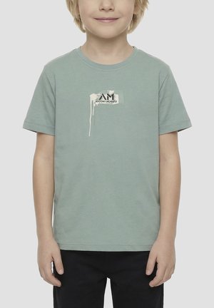 Niño con camiseta de manga corta verde menta con diseño de goteo de pintura blanca y pequeño logo de texto negro, combinado con pantalones negros.