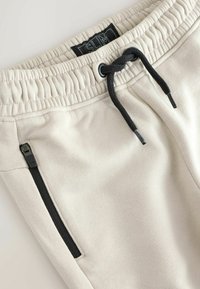 Light beige sweatpants with an elastic waistband, black drawstring, and zippered side pockets for secure storage. Soft textured fabric.