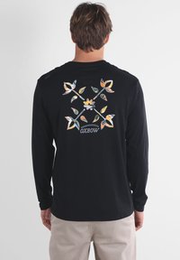 Chemise noire à manches longues avec un motif floral orange, jaune et vert au dos, avec le texte "OXBOW" sous le graphisme.