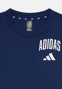 Camiseta deportiva de color azul marino con cuello redondo, logo blanco de "ADIDAS" y tres rayas blancas. Textura suave, manga corta.