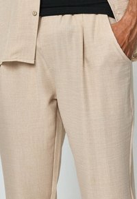Pantalon beige en tissu texturé, offrant une coupe décontractée, des poches latérales et un subtil design plié à l'avant. Accompagné d'une chemise légère.