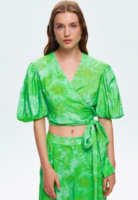 Groene cropped wikkeltop met poffende mouwen, voorzien van een gestructureerd bloemenpatroon en een strikdetail bij de taille; gecombineerd met bijpassende groene broek.